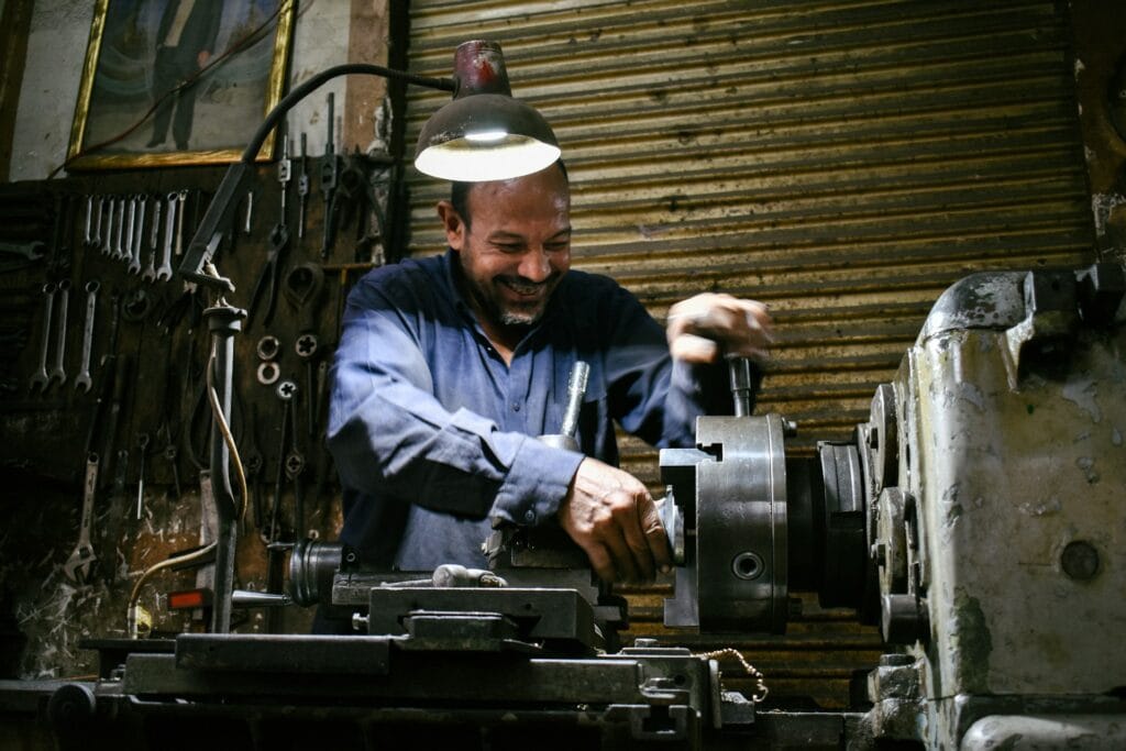 A machinist working at a lathe, with a rack of spanners on the wall and a bench lamp