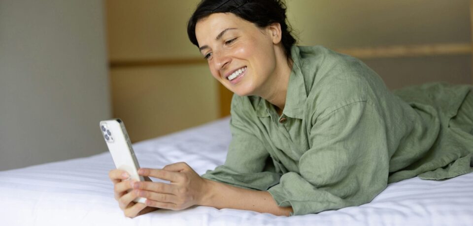 Cliente souriante allongée sur un lit d’hôtel, qui consulte son téléphone