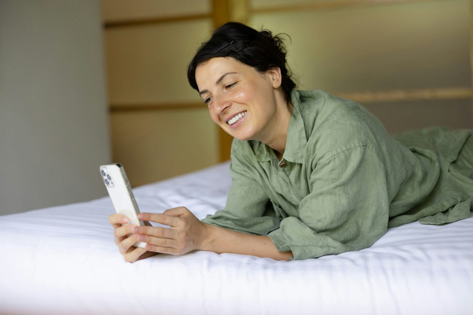 Cliente souriante allongée sur un lit d’hôtel, qui consulte son téléphone