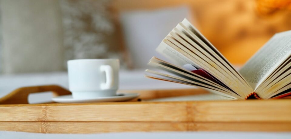 Livre ouvert et tasse blanche posés sur un plateau en bois, sur un lit