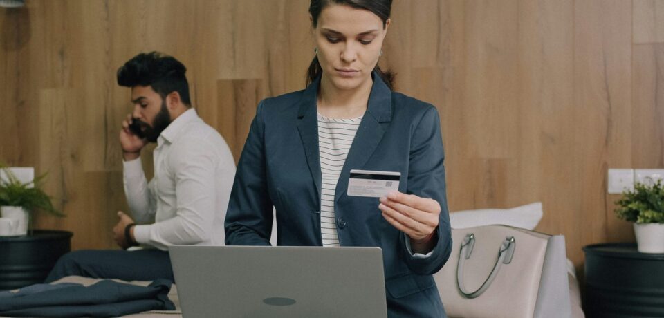Cliente en tenue de travail assise sur un lit d’hôtel, qui réserve sur son ordinateur portable, carte bancaire en main