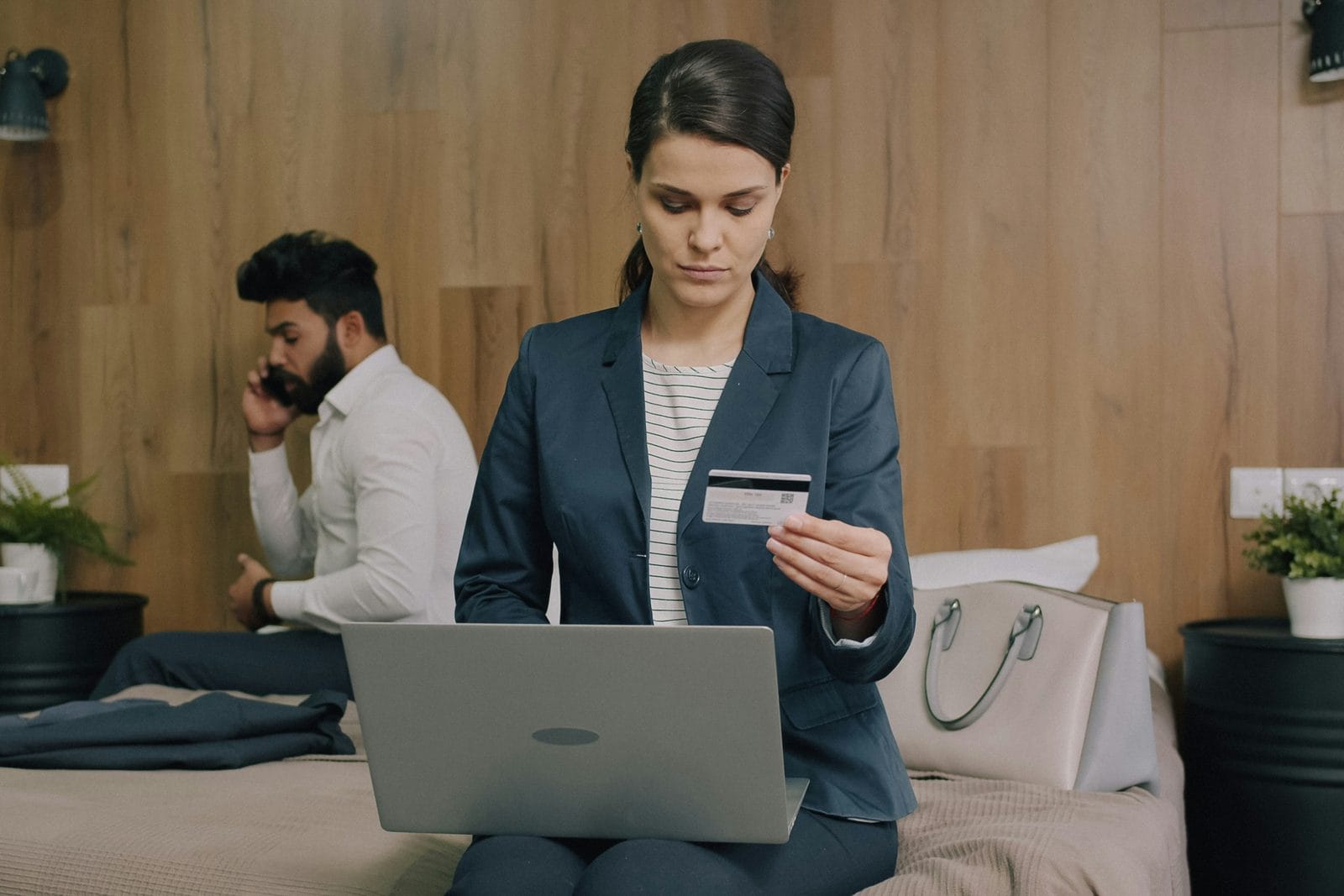 Cliente en tenue de travail assise sur un lit d’hôtel, qui réserve sur son ordinateur portable, carte bancaire en main